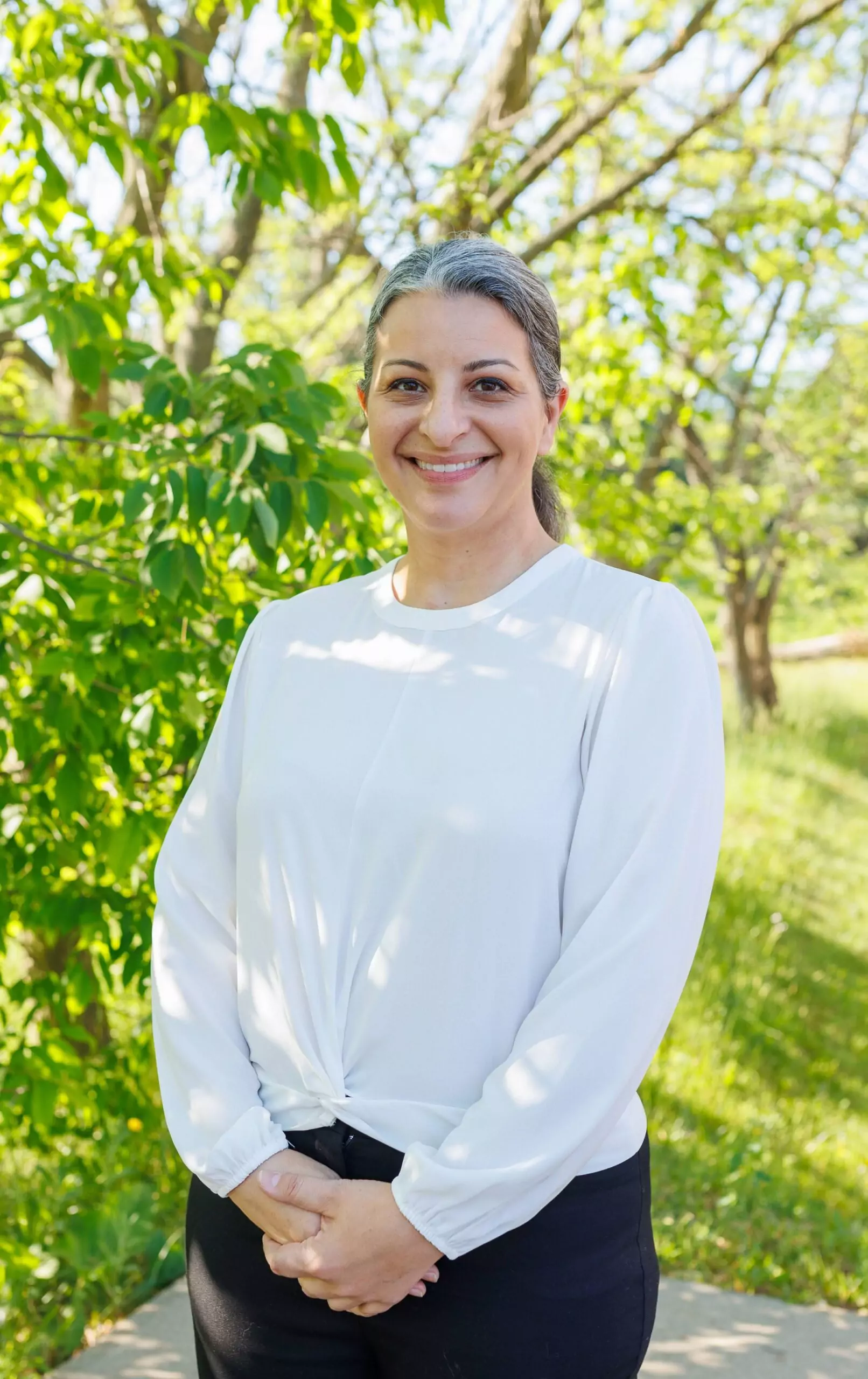 Mary Riposo standing in front of a green tree and grass. She has salt-and-pepper hair pulled back and is smiling. She is wearing a white shirt and dark pants.