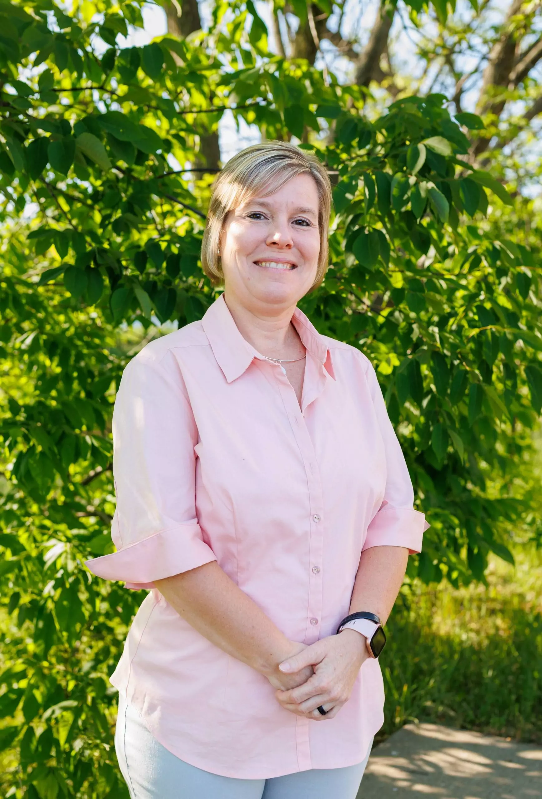 Angela Rodgers standing in front of a green tree. She has short blond hair and is smiling with her arms folded in front of her. She is wearing a pink button up shirt and tan pants.