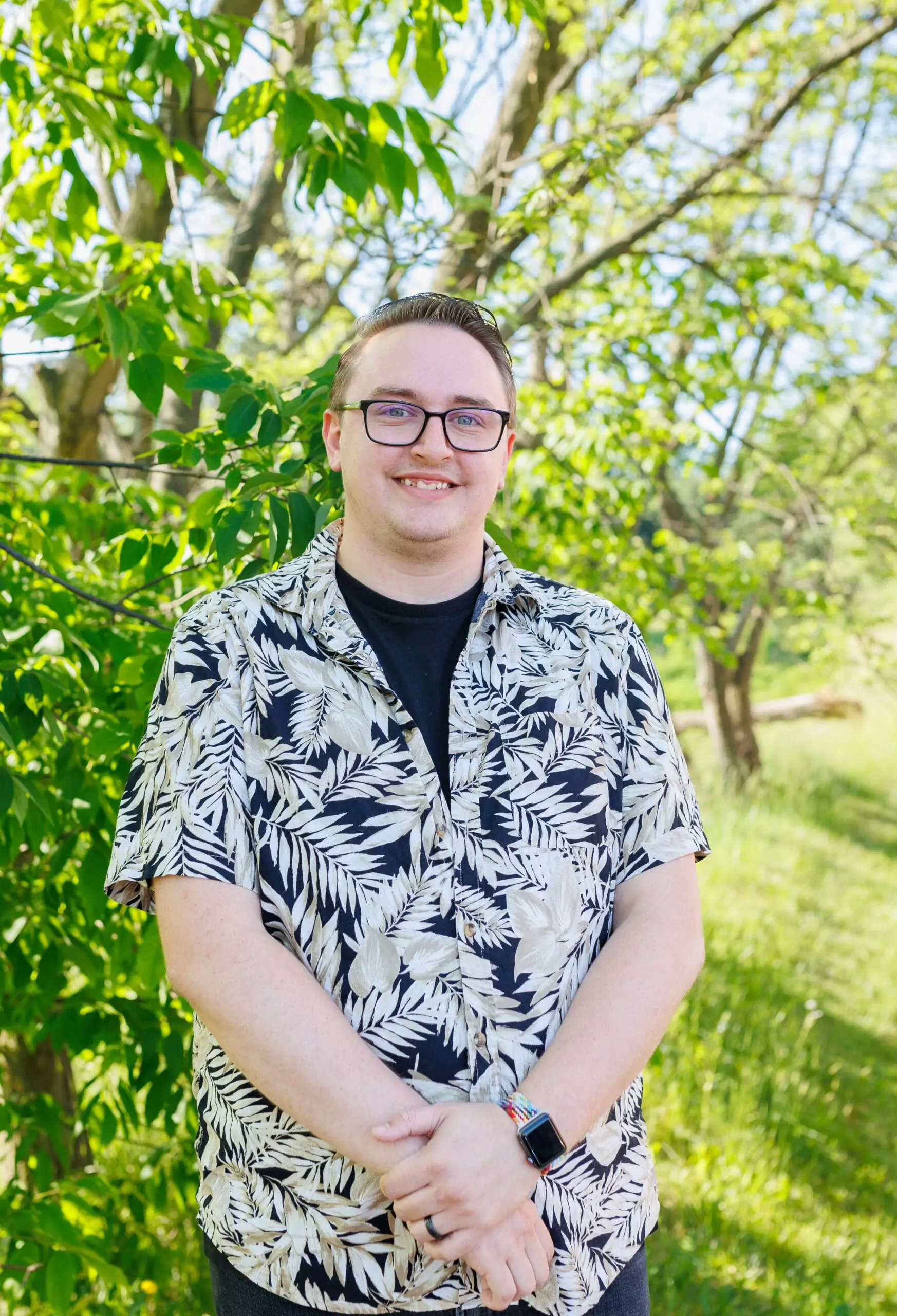 Jared Sparks standing in front of a green tree. He has short dark hair and round, dark glasses. He is wearing a black and white aloha shirt over a black t-shirt.
