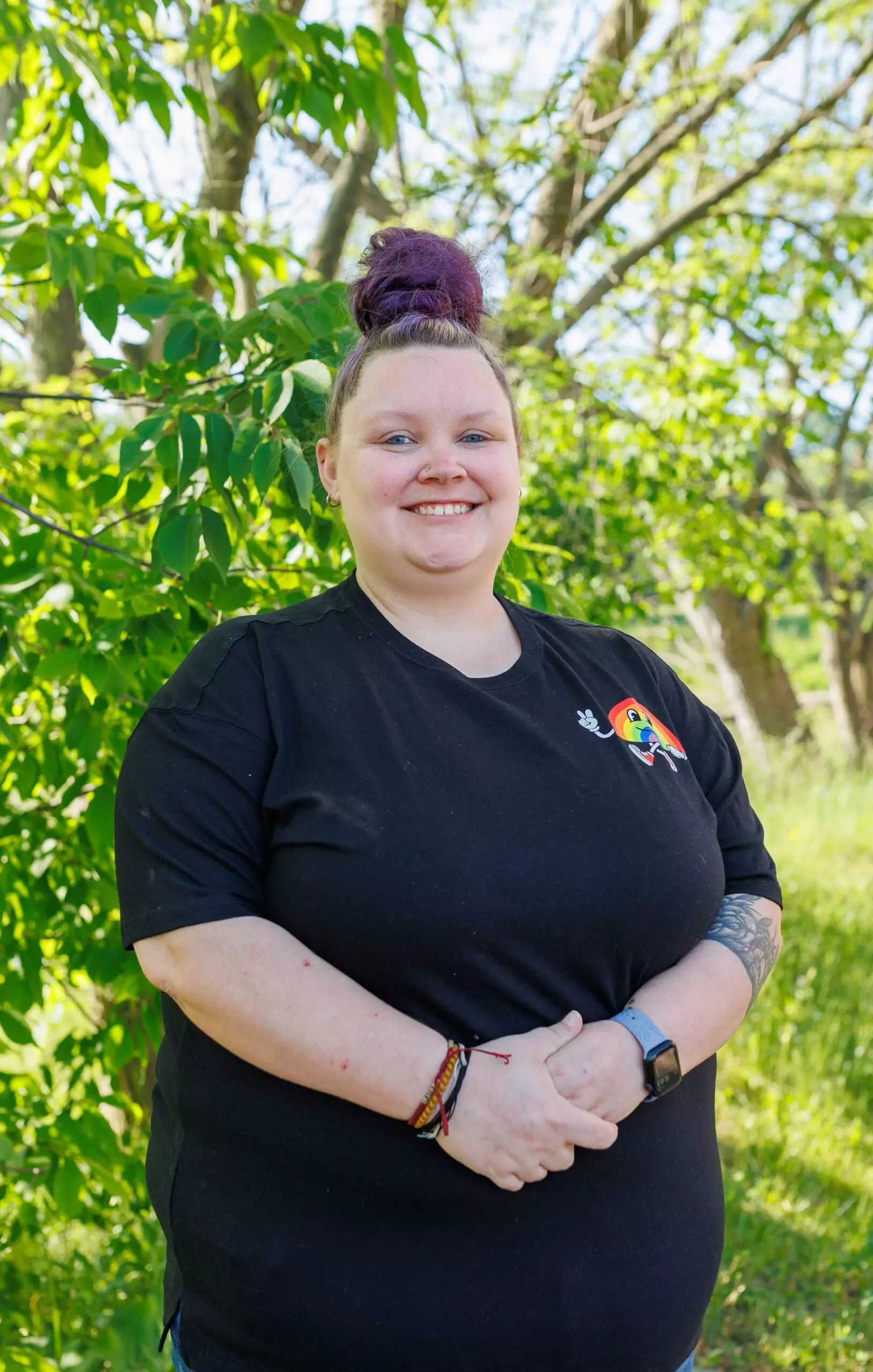 Taylour Kidd standing in front of a tree, outside. She is a white female and smiling with her hair up in a bun. She is wearing a short-sleeve black shirt with a animated rainbow giving a thumbs up on the left-chest.