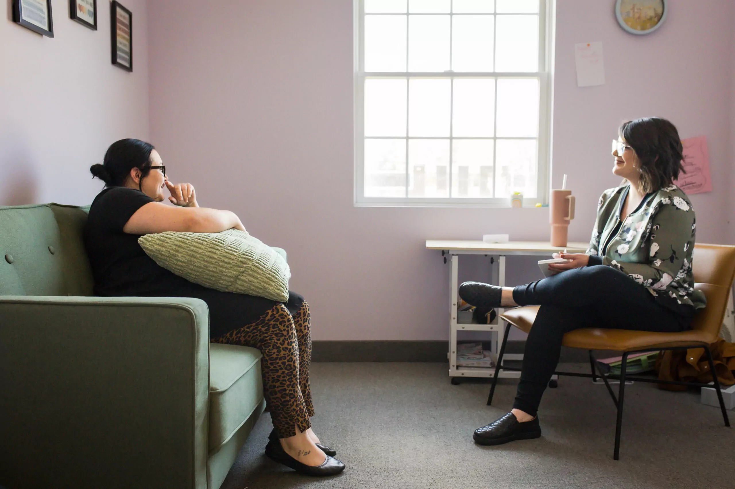 A female therapist sitting at right talks with a female client sitting on a couch at left. A window with sunlight is in the wall behind them.