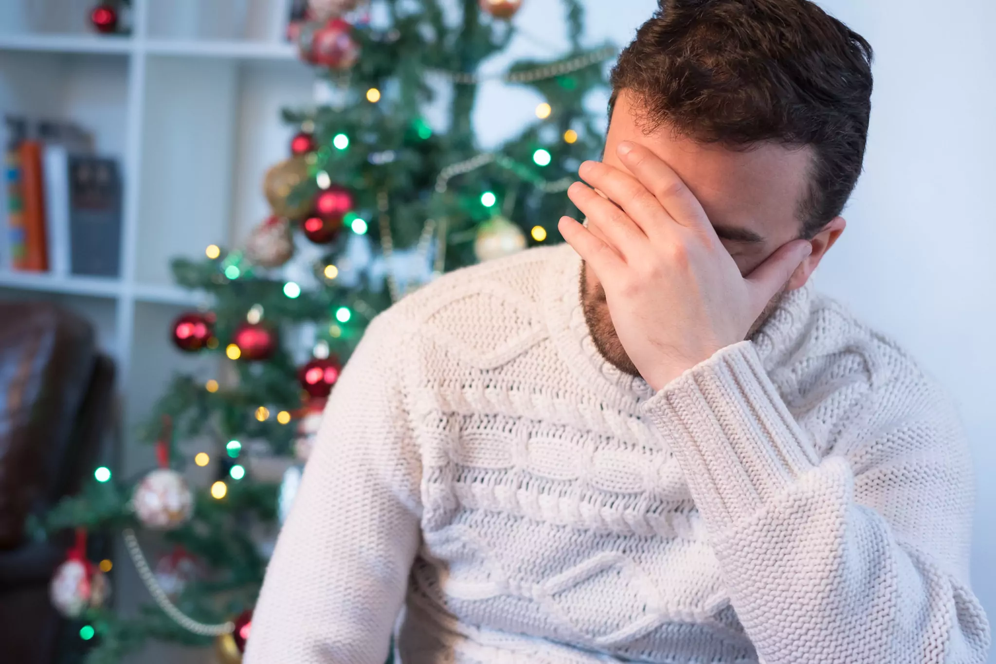 Man feeling depressed, wearing a white sweater and holding his head in his hand while sitting in front of a Christmas tree.