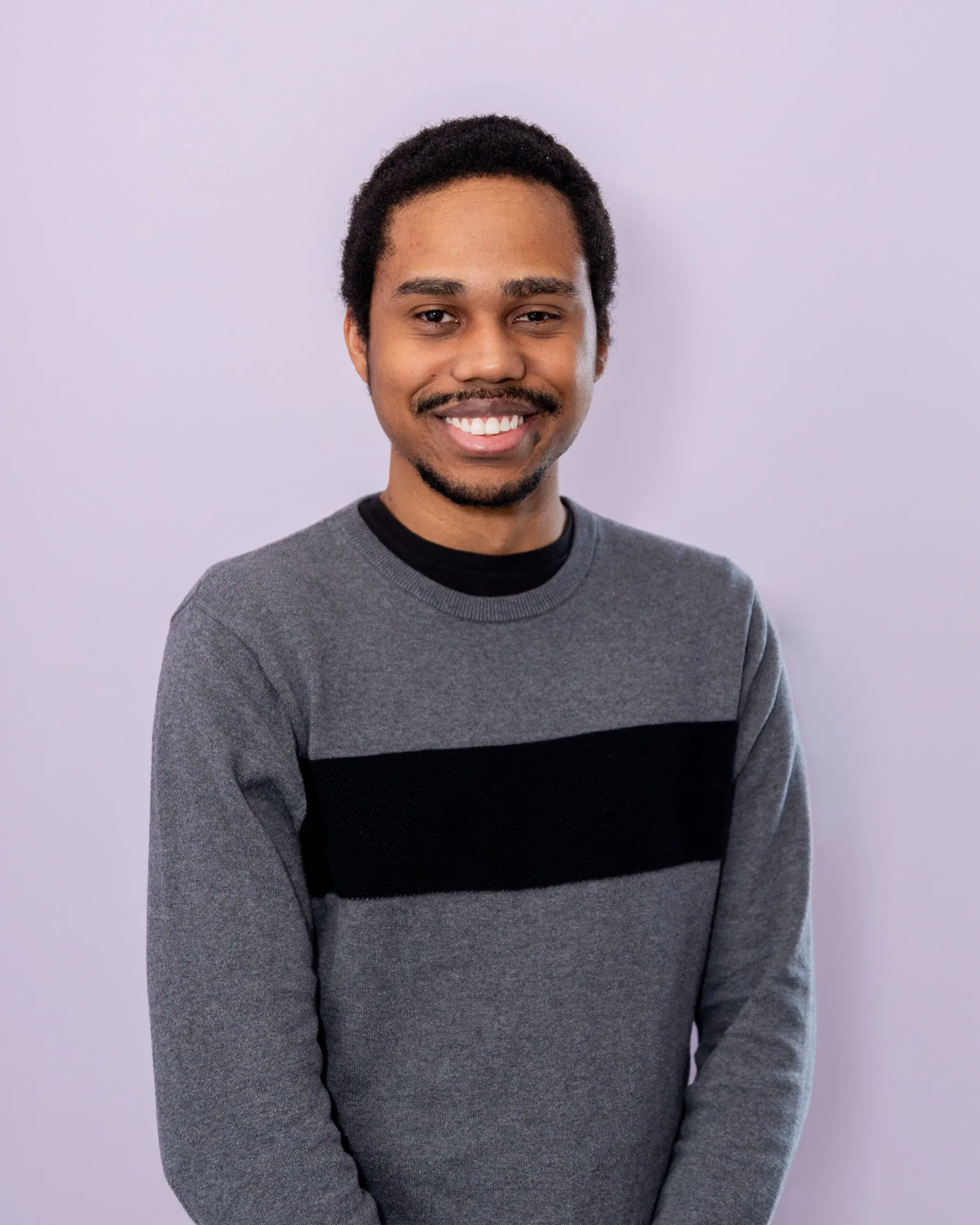 Black male, smiling and wearing a grey sweater with a black stripe. Thomas is standing against a purple wall.