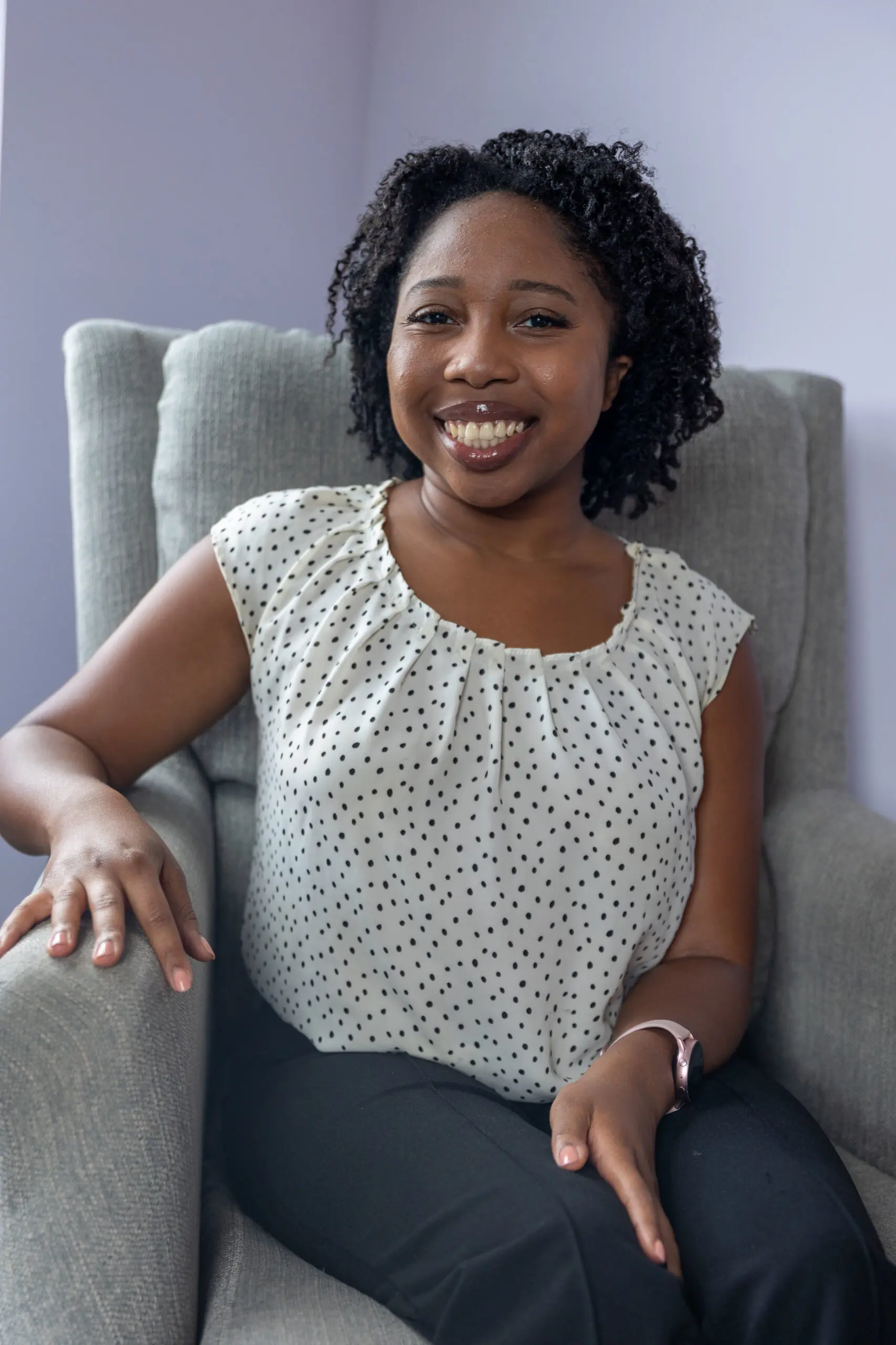 A black woman with medium length dark hair, smiling, and siting. Rachel is wearing a white sleeveless shirt with dots and dark pants.