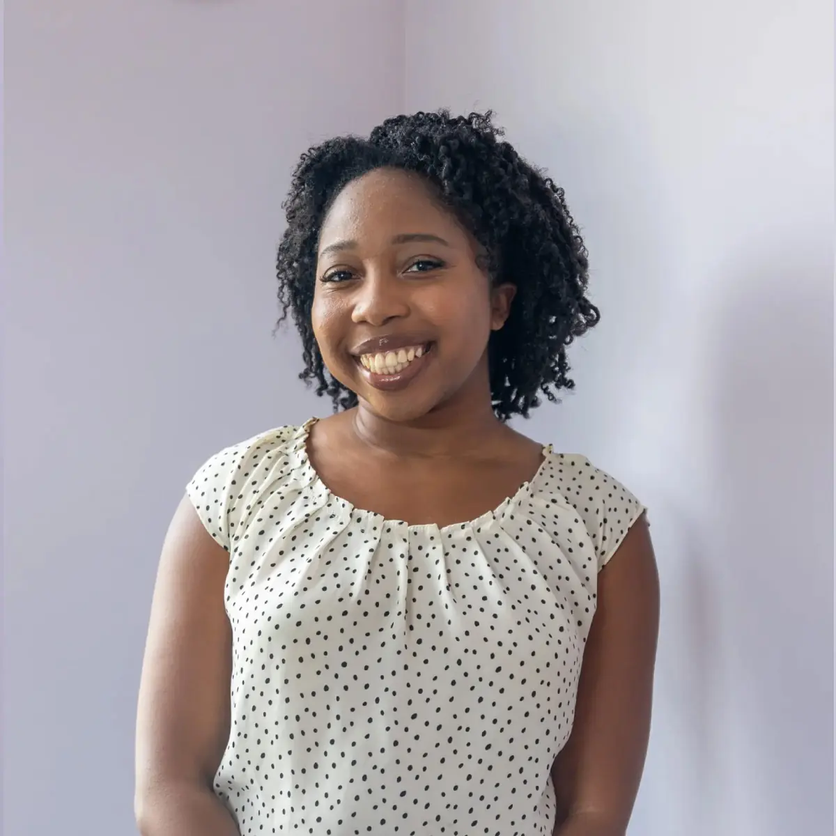 A black woman with medium length dark hair and smiling against a purple wall. Rachel is wearing a white sleeveless shirt with dots and dark pants.